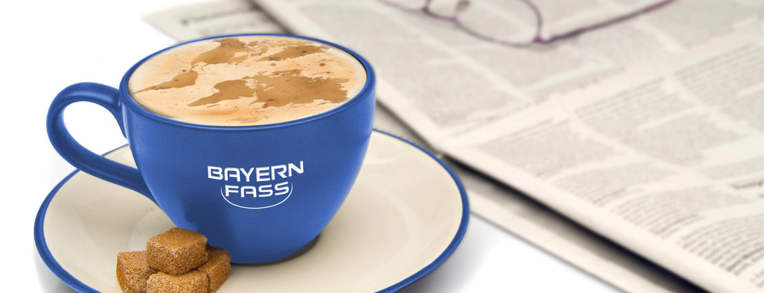 eine blaue Kaffeetasse mit weißem Bayern-Fass Logo steht auf einer Untertasse, daneben eine Tageszeitung